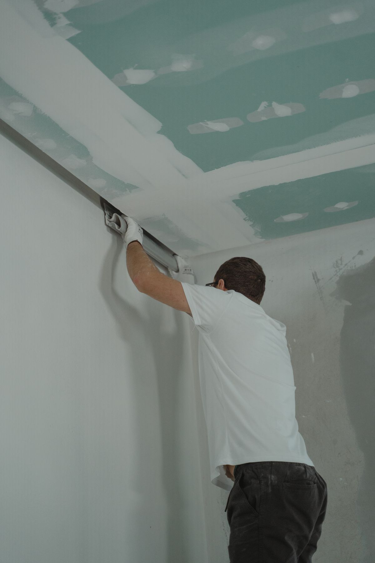 Worker smoothing joint compound along the wall-to-ceiling corner beneath new green drywall