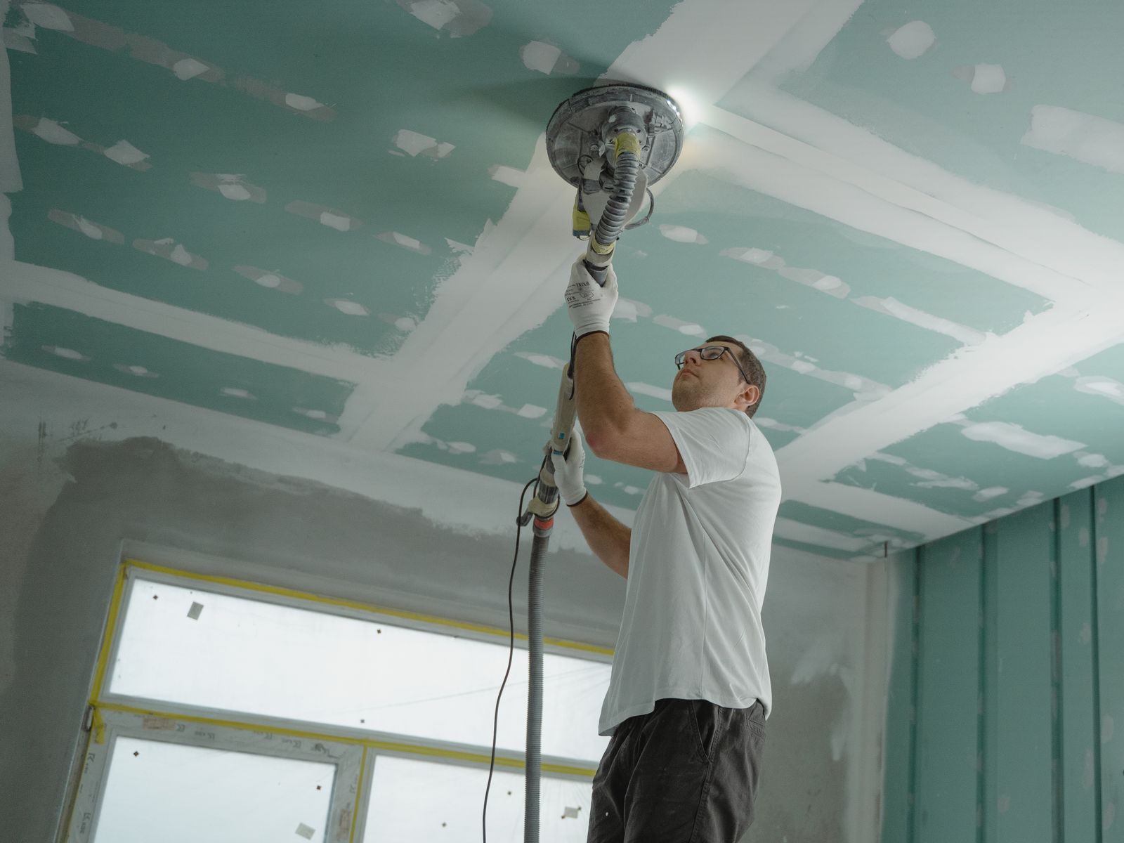 Worker using a dust-extracting drywall sander on a freshly taped ceiling