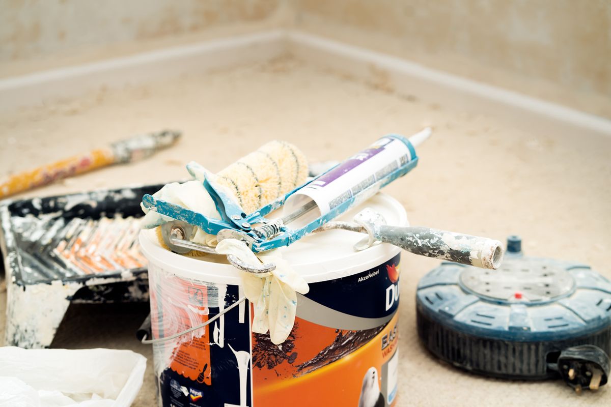 Paint bucket topped with a roller sleeve, caulk gun, and used brushes beside a paint tray on a drop-clothed floor